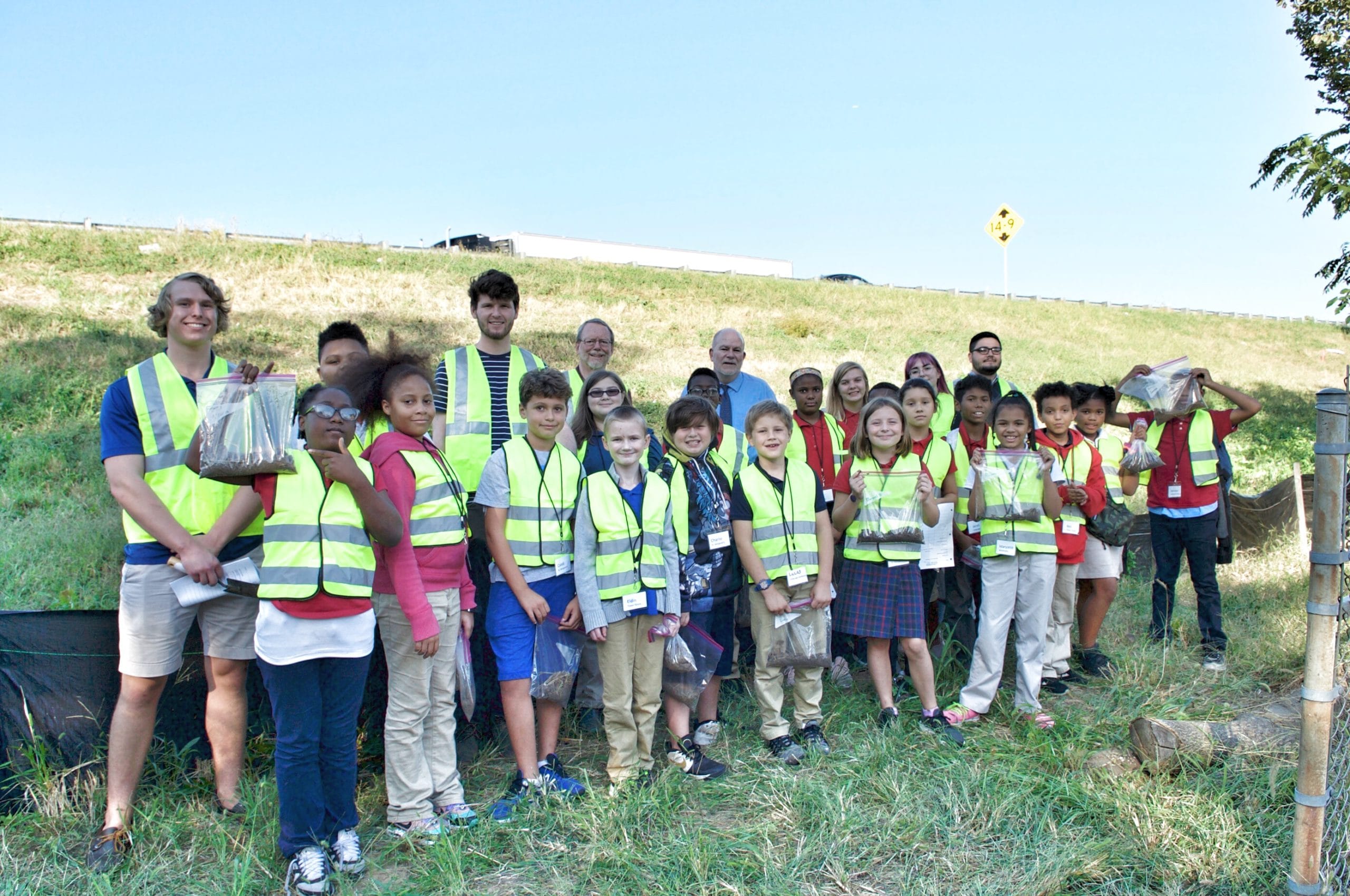 A group of students in yellow safety vests pose for the camera. The students are outdoors and holding ziploc bags of soil.